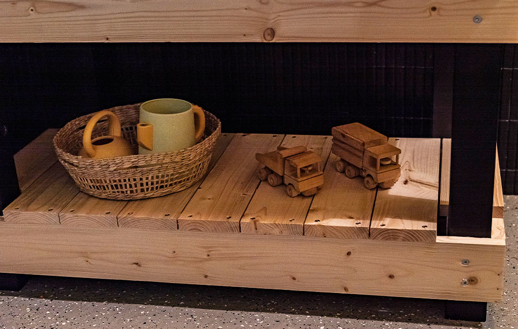 The lower section of the mud kitchen holds toy trucks and a basket with a watering can and cups.