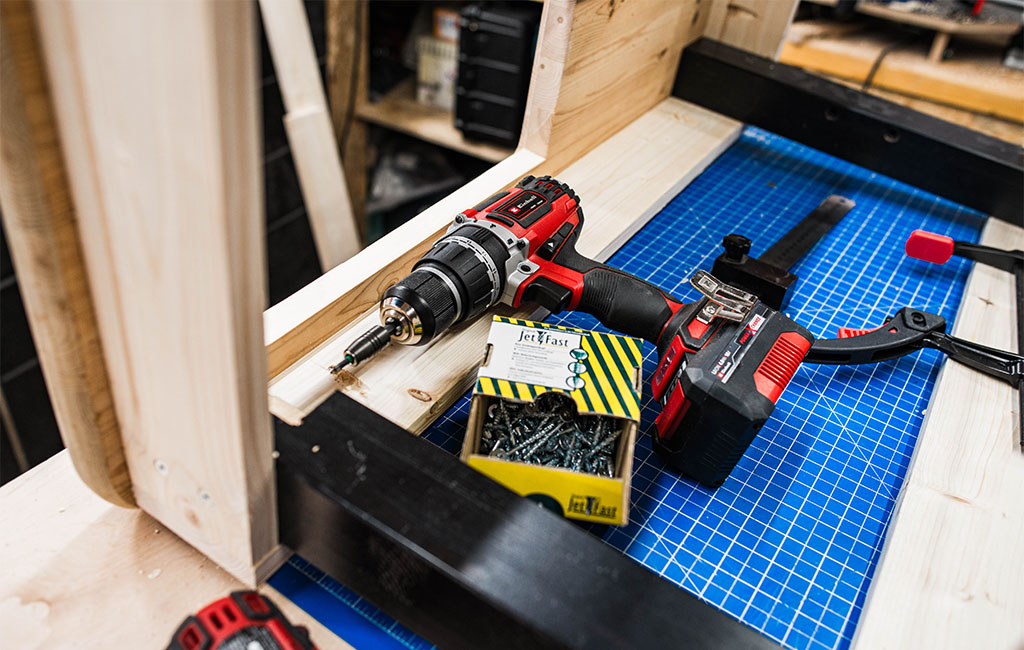 Cordless screwdriver, box of screws and clamp lie on the wooden frame of the mud kitchen.