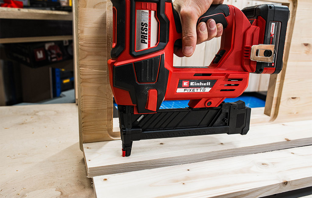 A hand holding a Einhell cordless nailer against a wooden part during mud kitchen assembly.
