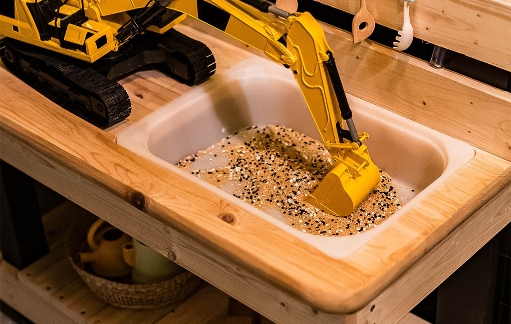 Close-up of the toy sink in the mud kitchen filled with sand, grains, and a yellow toy excavator.