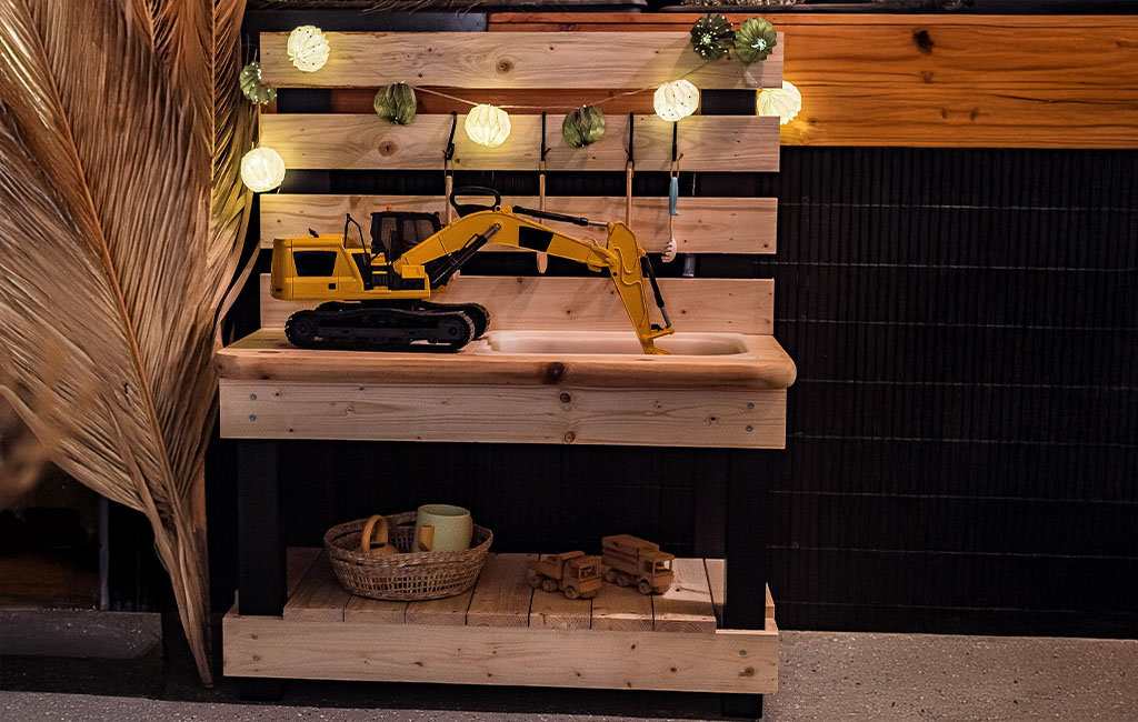 Fully assembled mud kitchen with toy excavator in the sink, wooden hooks, play dishes, and wooden toy trucks.
