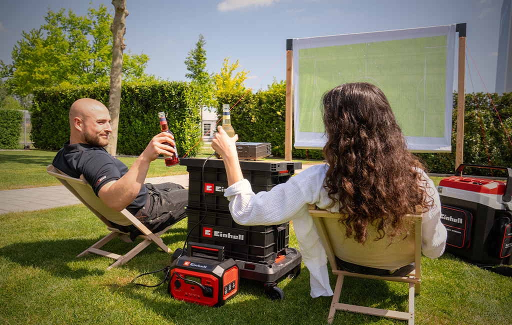 Ein Mann und eine Frau sitzen in Liegestühlen auf dem Rasen, stoßen mit Flaschen an und schauen Fußball auf der DIY-Leinwand.