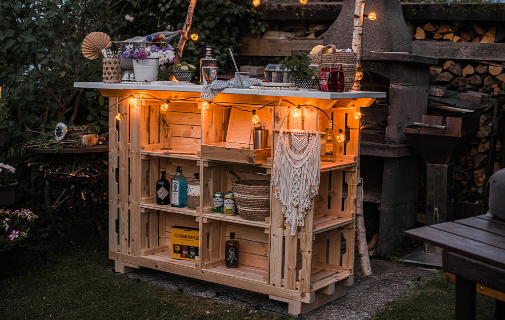DIY garden bar made from wooden crates, decorated with string lights, plants, and drinks in a cozy evening setting.