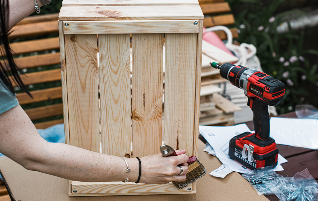 A woman brushes a wooden crate while an Einhell cordless drill rests on the table next to her.
