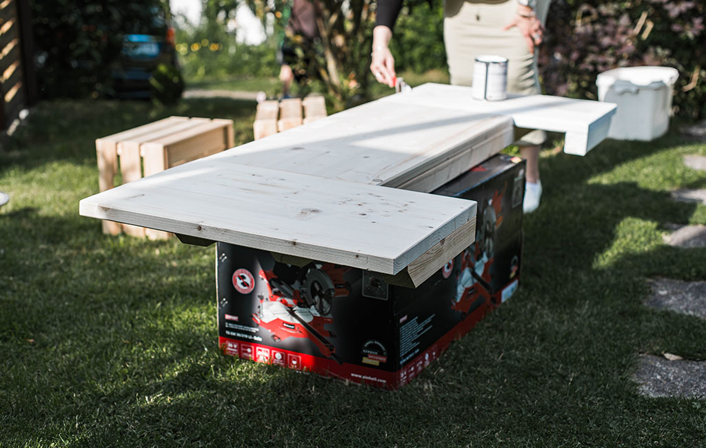 A woman paints the white bar top resting on cardboard boxes in a garden setting.