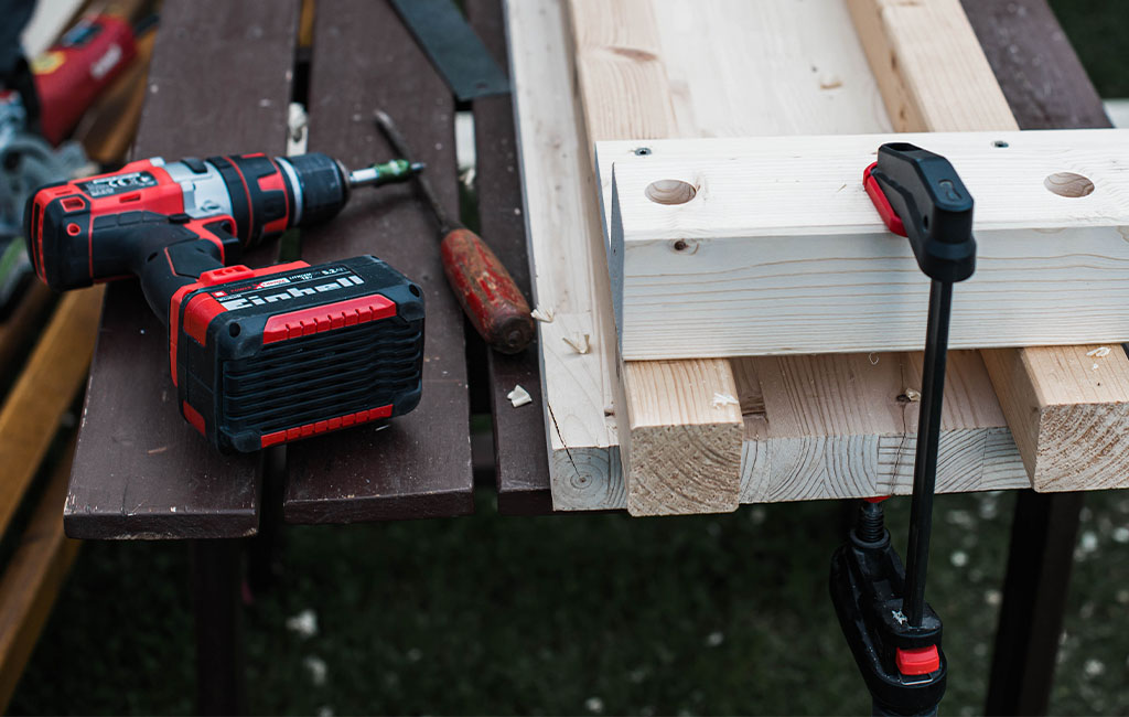 An Einhell cordless drill rests on a table beside clamped wooden parts and a clamp.