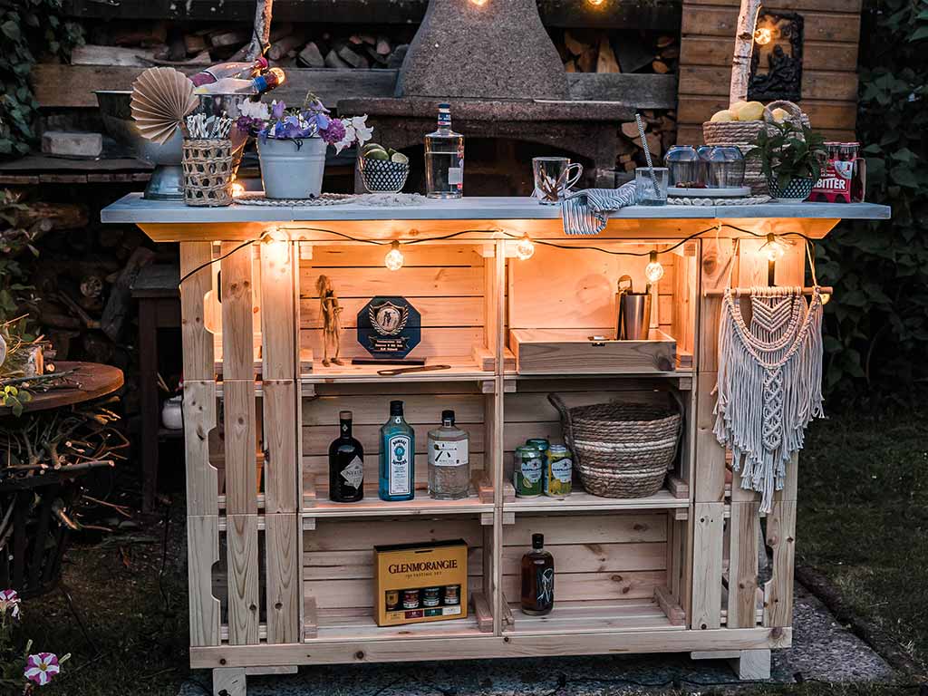 DIY garden bar made from wooden crates, decorated with string lights, plants, and drinks in a cozy evening setting.