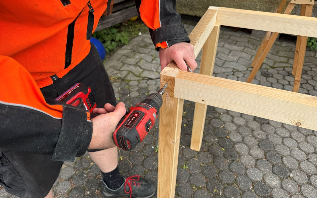 Eine Person befestigt ein Kantholz mit Schrauben am Untergestell des Pflanztisches.