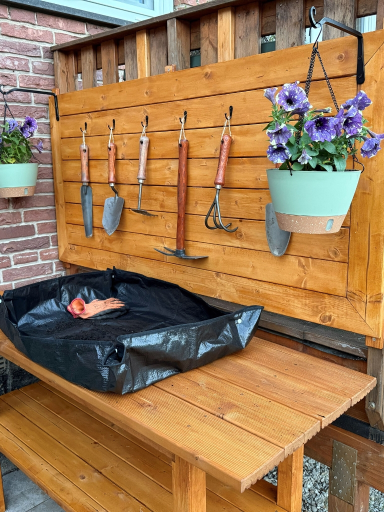 Ein fertig dekorierter Pflanztisch mit Rückwand, Gartenwerkzeugen, Blumenampel und Pflanzunterlage.