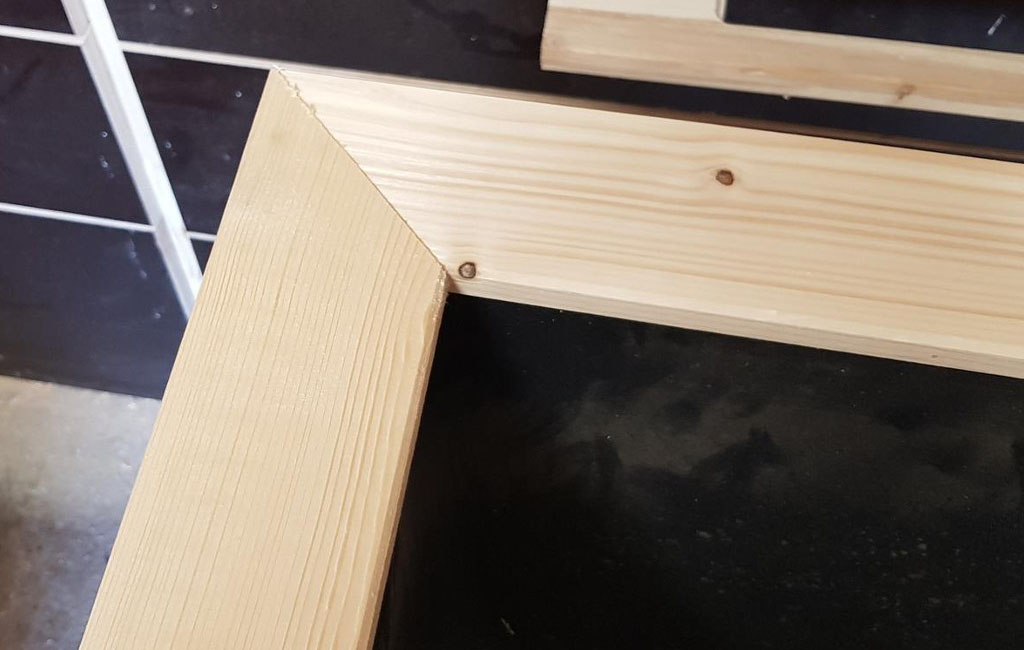 Nahaufnahme eines hellen Holzrahmens mit Gehrungsschnitt an der Ecke, auf einem schwarzen Untergrund.