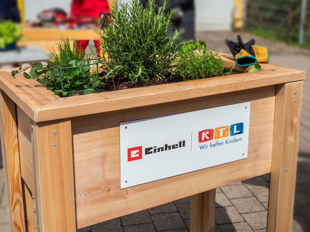 Ein Hochbeet aus Holz mit Kräutern bepflanzt, auf der Vorderseite ein Schild mit den Logos von Einhell und RTL.