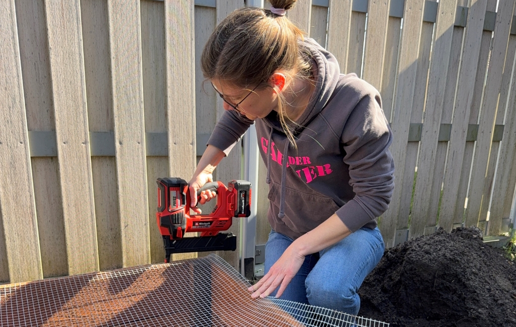 Eine Frau kniet im Garten und befestigt mit einem Einhell Akku-Nagler ein Drahtgitter am Boden eines Holzrahmens.