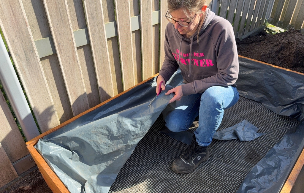 Eine Frau kniet in einem Holzrahmen und schneidet graue Teichfolie mit einer Schere passend zu.