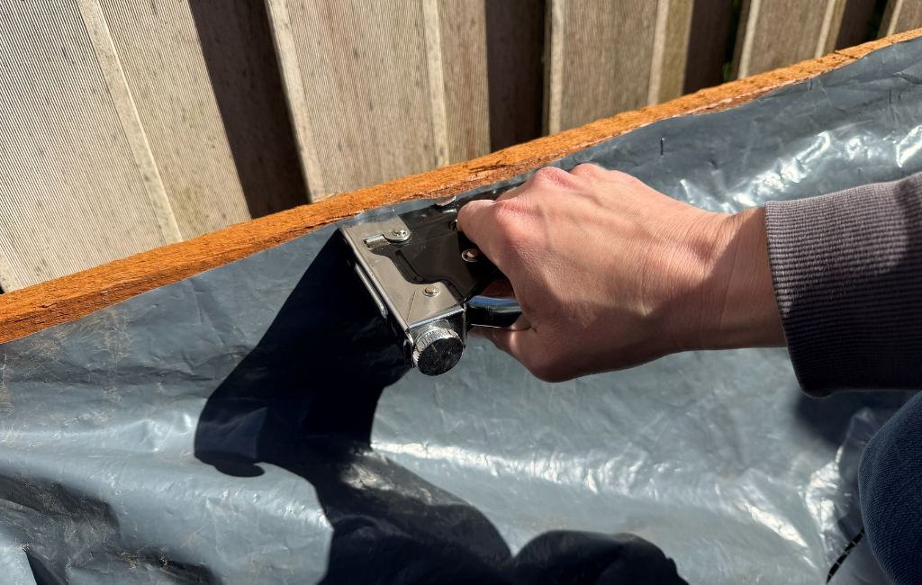 Eine Hand befestigt mit einem silbernen Handtacker die graue Teichfolie am inneren Rand eines Holzrahmens.