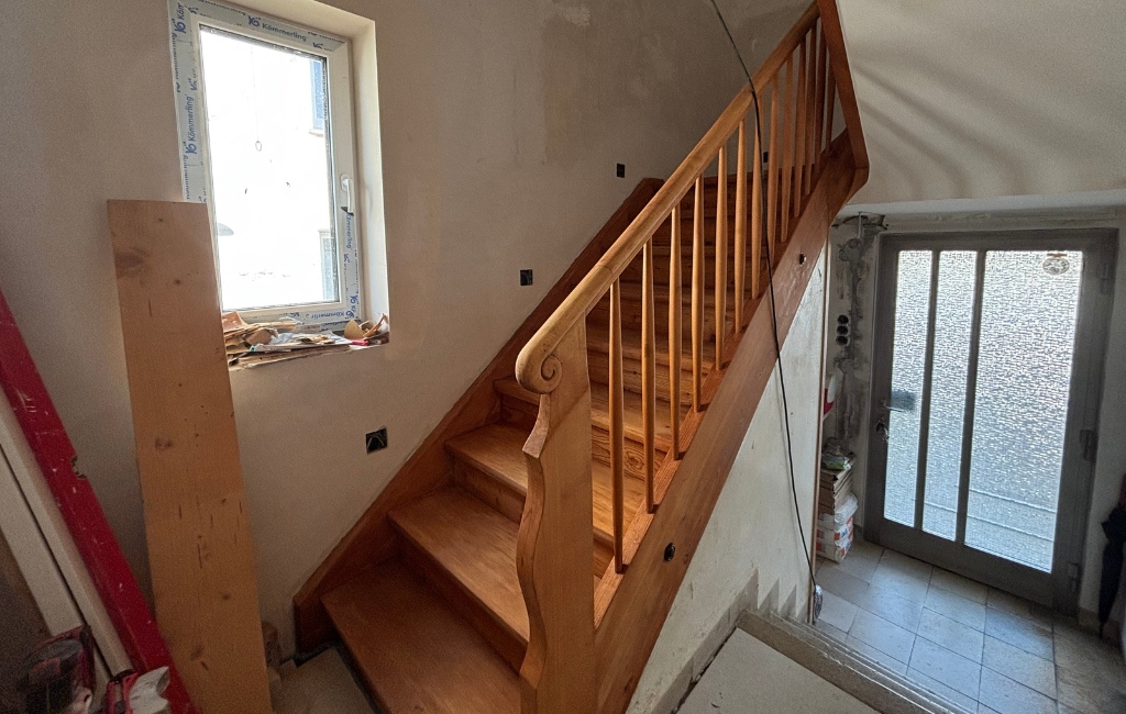 Holztreppe mit Geländer in einem renovierten Flur, links ein Fenster, unten eine Glastür.