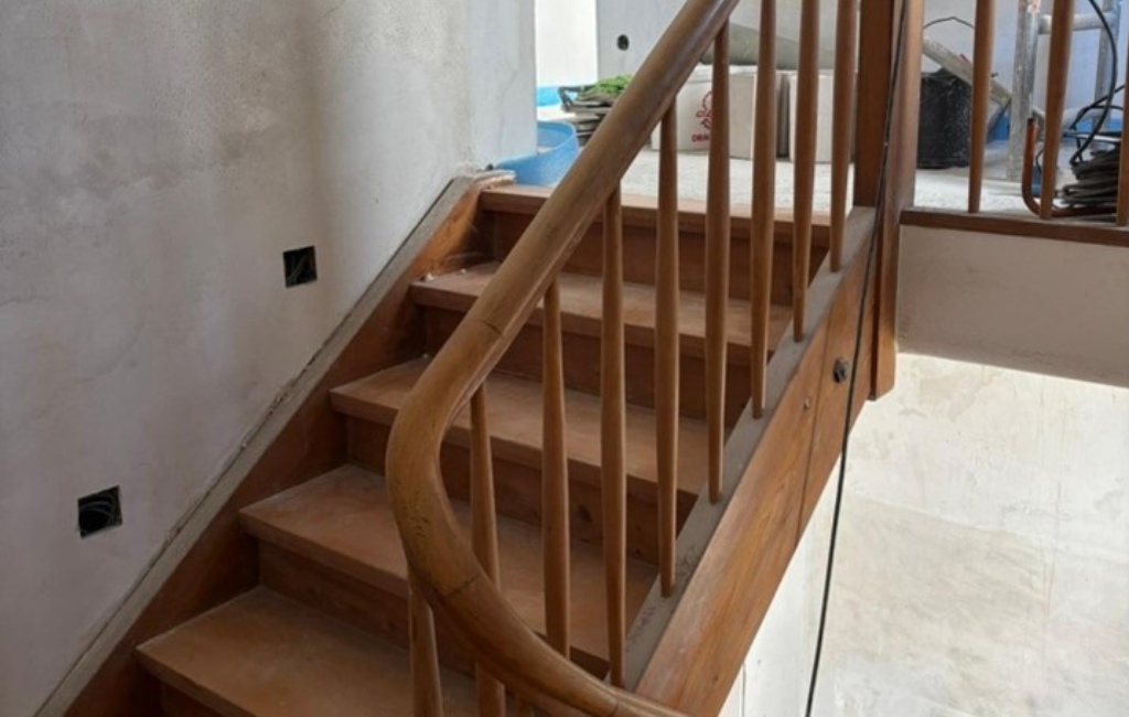 Holztreppe mit Geländer in einem renovierten Flur, links ein Fenster, unten eine Glastür.