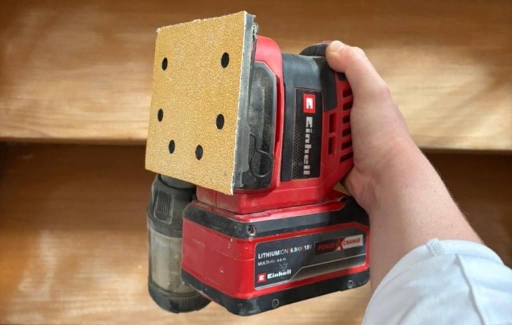 Eine Hand hält einen roten Einhell Akku-Schwingschleifer mit eingesetztem Power X-Change Akku.