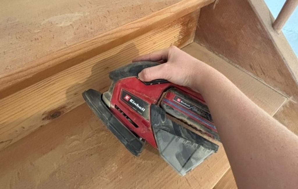 Eine Hand schleift eine Holzstufe mit einem roten Einhell Multischleifer.