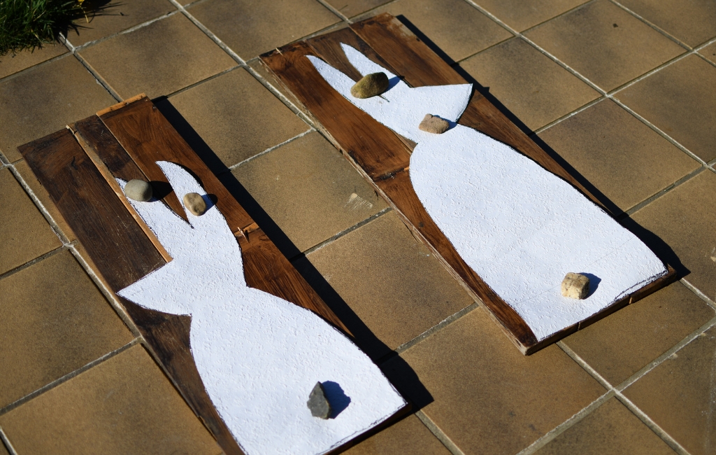 Holzplatten mit weiß aufgemalten Hasensilhouetten liegen beschwert mit Steinen auf Fliesen.
