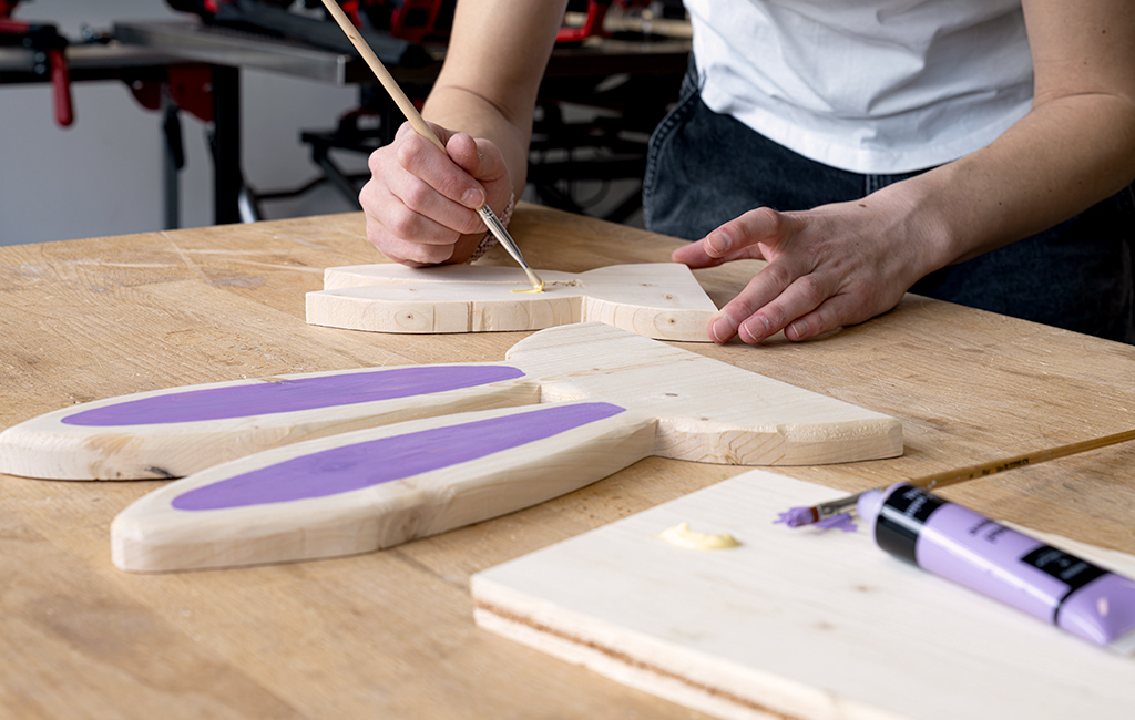 Eine Person bemalt die Ohren einer Holzhasenform mit einem Pinsel in lila Farbe.