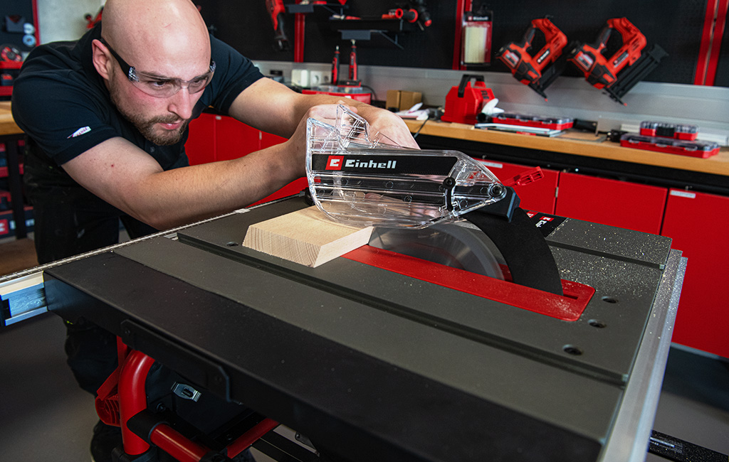 A man cuts a wooden piece using an Einhell table saw, with the blade covered by a safety guard.