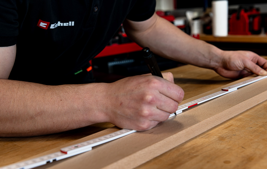 A man measures a wooden board with a folding ruler and marks a spot using a black marker.