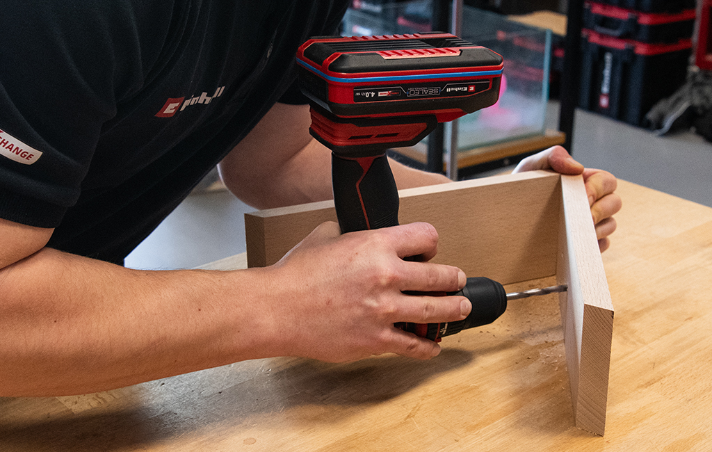 A man drills a hole into two wooden slats positioned at an angle using an Einhell cordless drill driver.