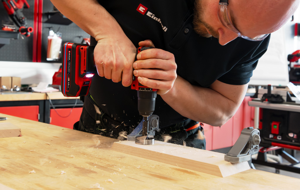 A man drills holes into a wooden piece clamped on a workbench using an Einhell cordless drill driver.