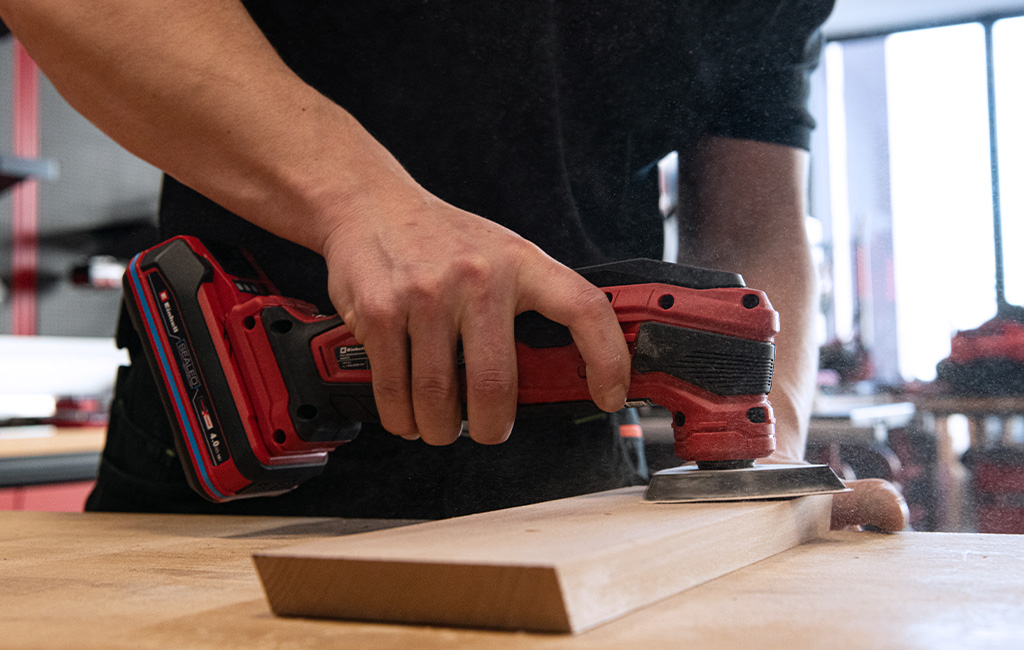 A man sands a wooden piece on a workbench using an Einhell cordless multitool.