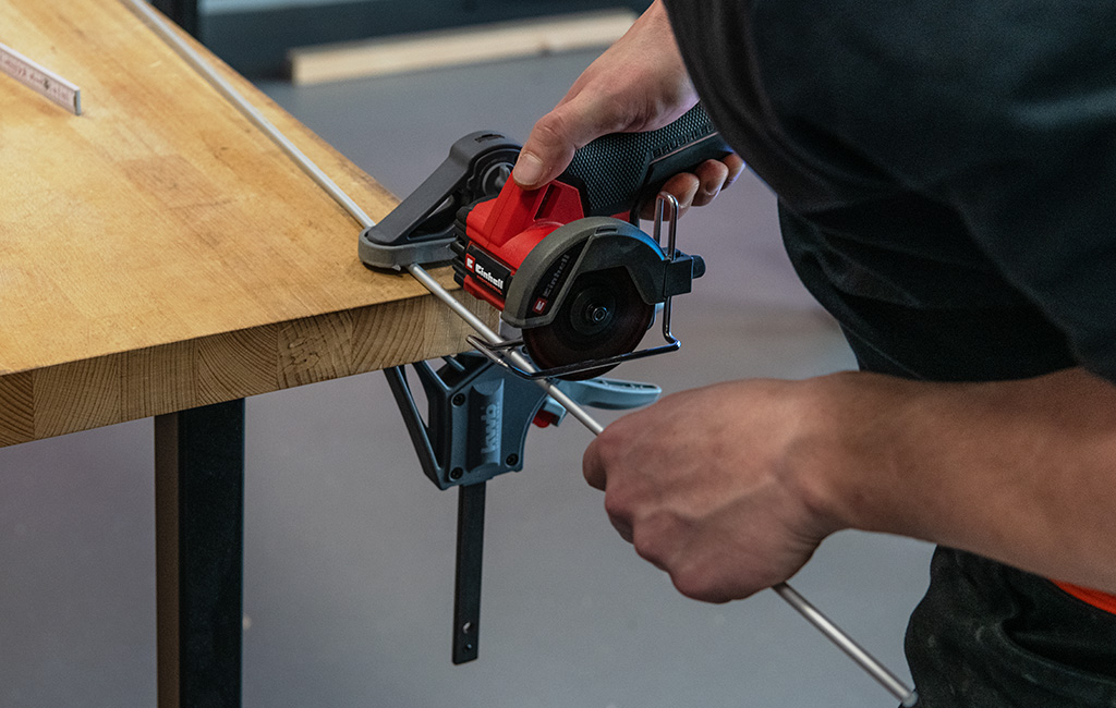 A man cuts a metal rod clamped to a workbench using an Einhell cordless cut-off tool.