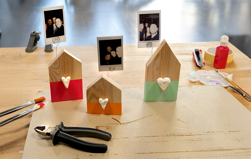 Three house-shaped wooden photo holders with colored bases and white hearts, arranged on a craft table.