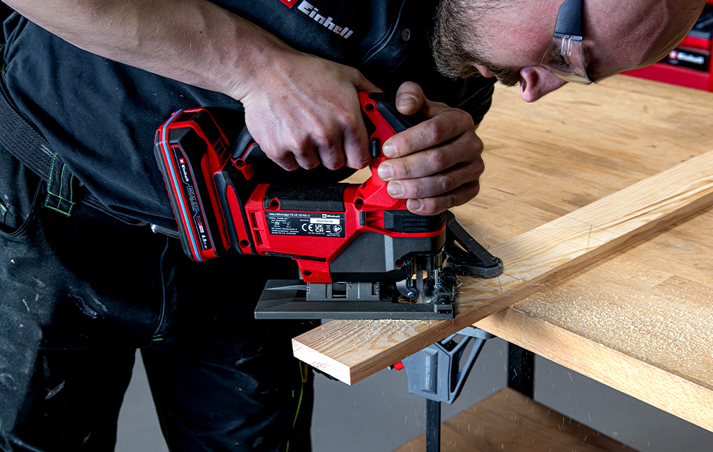 A person is cutting a house shape out of a wooden board using an Einhell cordless jigsaw.