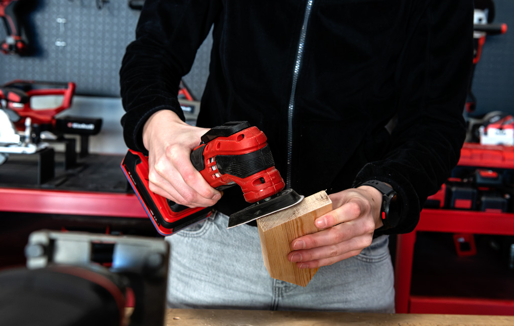 A person is sanding a small wooden house with a red Einhell cordless sander.