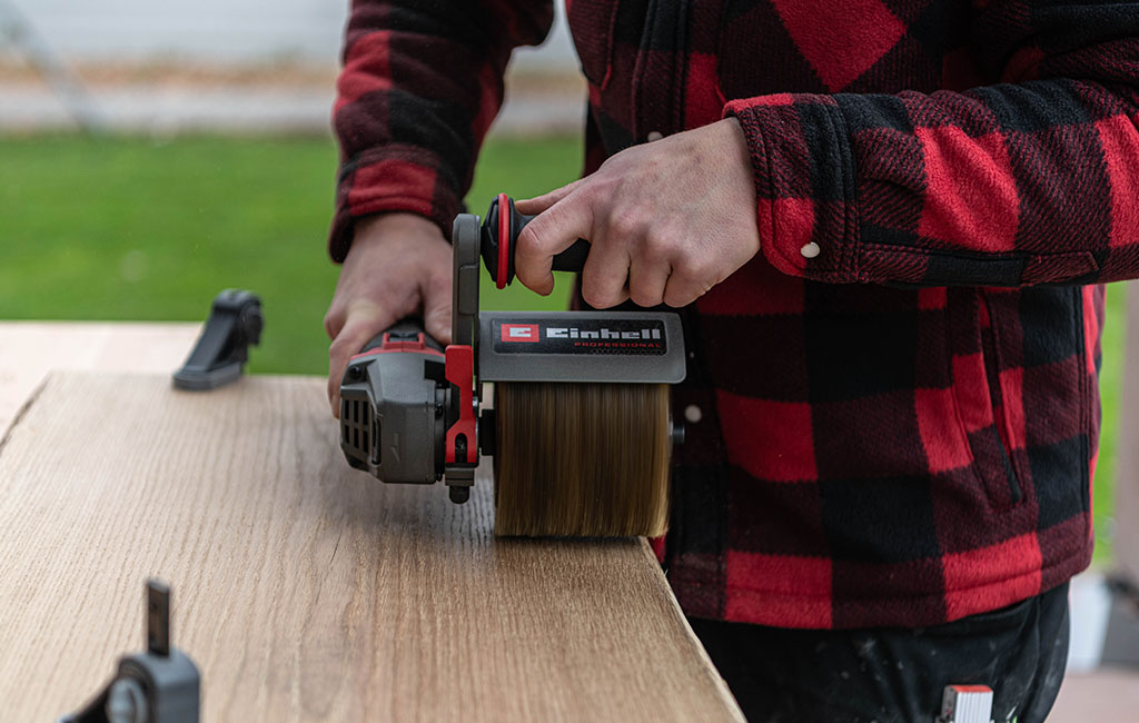 Eine Person bürstet die Oberfläche einer Holzplatte mit einer Akku-Bürstenschleifmaschine von Einhell.