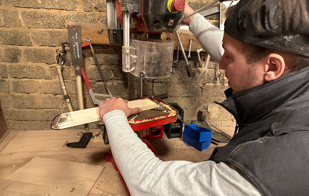 Person drilling into a wooden board with a bench drill in a workshop, holding the wood on the drill table during machining.