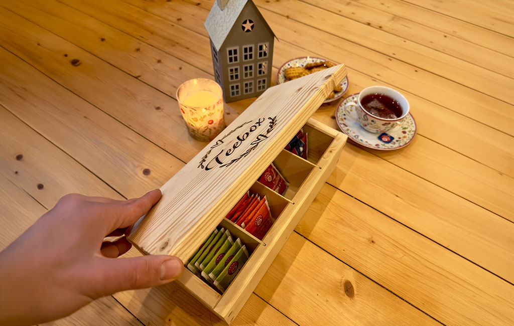 Handmade wooden tea box with engraved “tea box” lid and sorted tea bags inside, placed on a wooden table next to a cup of tea and candle.