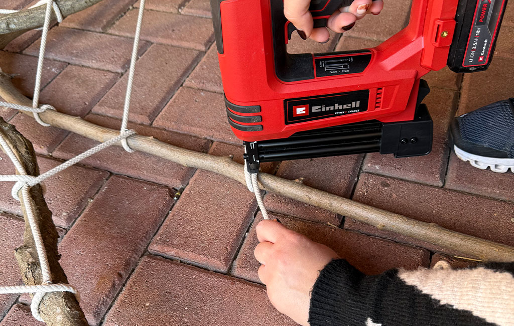 A person fastens a rope to a branch on the ground using an Einhell cordless stapler.