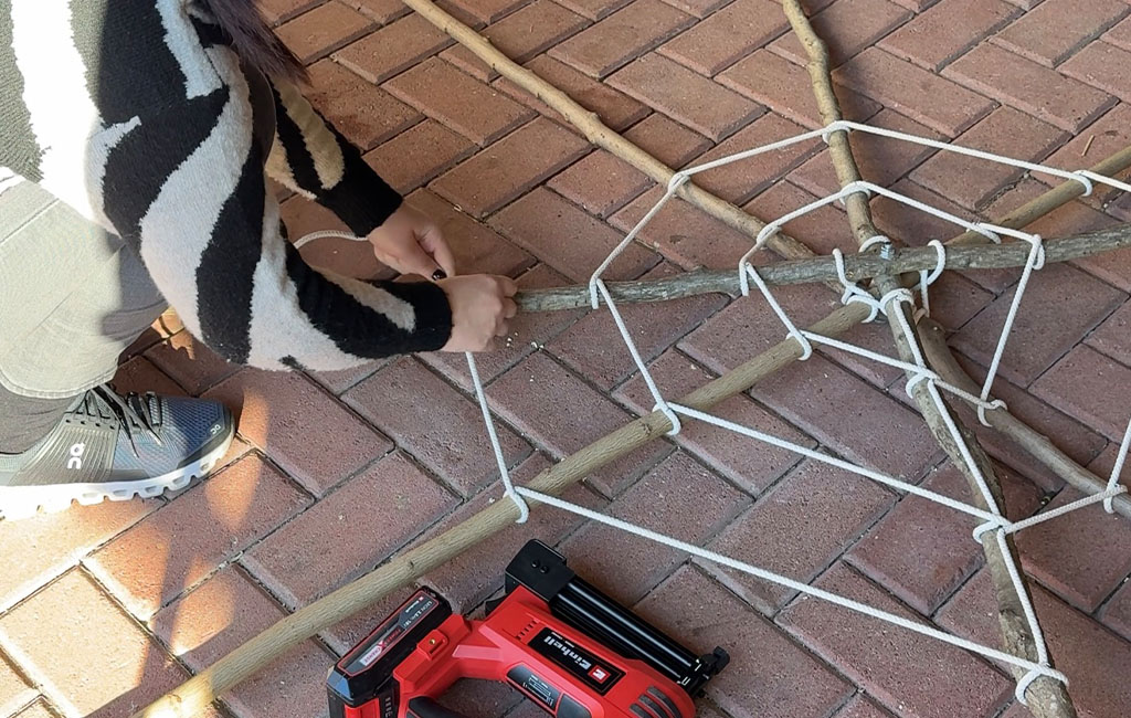 A person ties a spider web from rope to a frame made of branches on the ground.