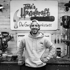Tobias Peters, wearing a hoodie and baseball cap, stands smiling in a workshop in front of the sign “Tobi’s Handcraft – Die Garagenwerkstatt”.