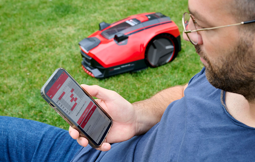 Ein Mann sitzt auf dem Rasen und steuert den Einhell Mähroboter FREELEXO CAM mit einer App auf dem Smartphone.