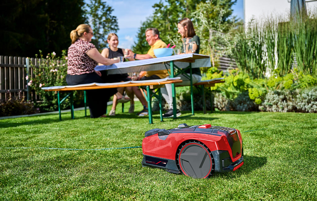 Der Einhell Mähroboter FREELEXO CAM mäht Rasen, während im Hintergrund vier Personen an einem Gartentisch sitzen.