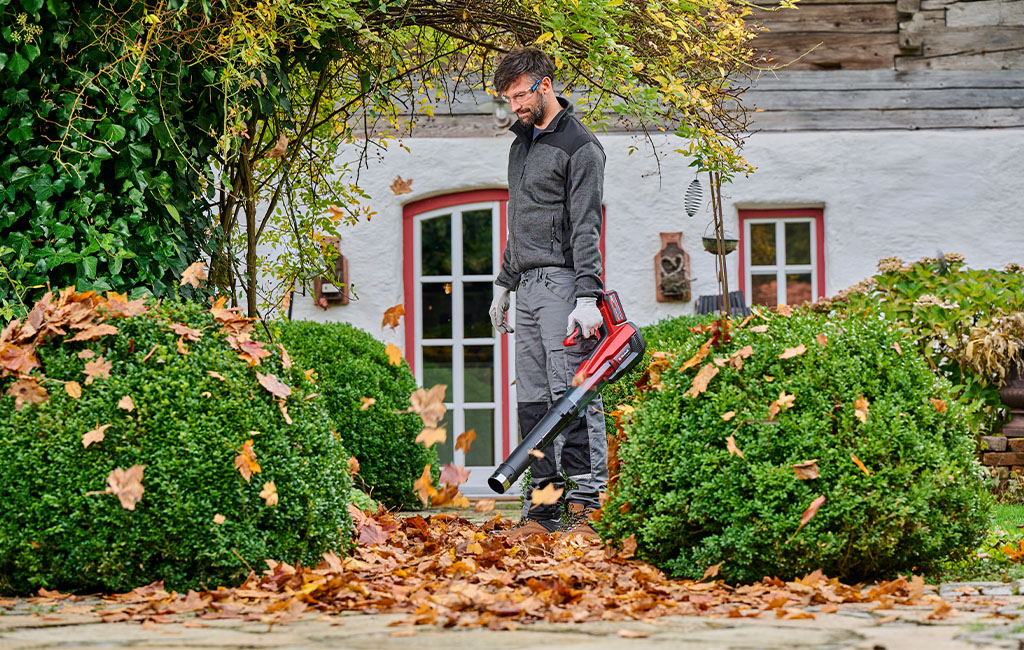 Ein Mann entfernt Herbstlaub mit einem Einhell Akku-Laubbläser vor einem Haus.
