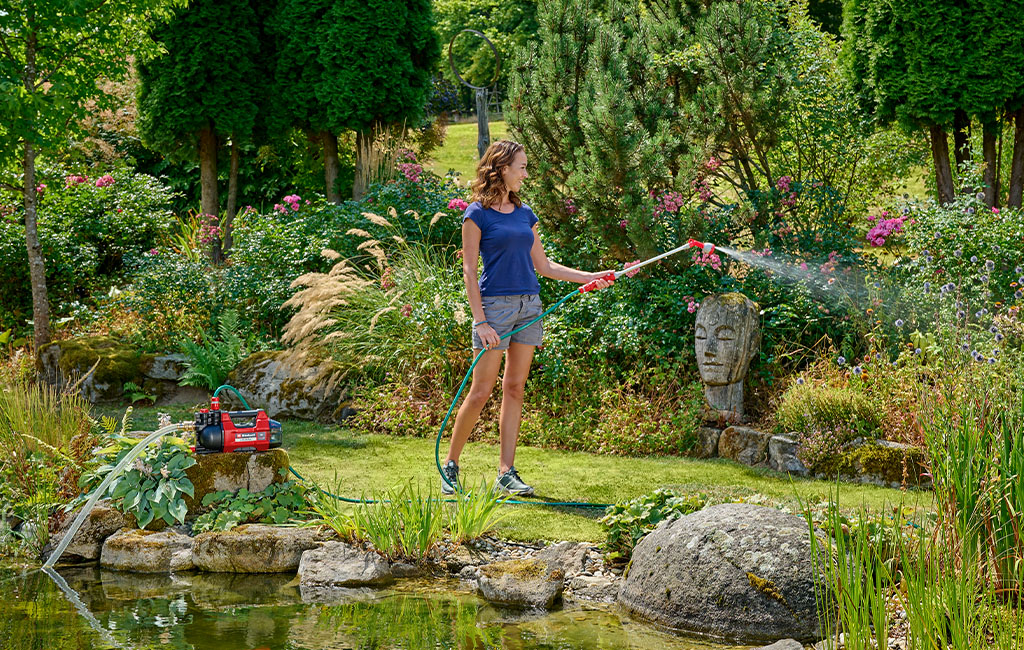 Eine Frau gießt Pflanzen mit einer Sprühlanze an einem Gartenteich, neben ihr eine Einhell Gartenpumpe.