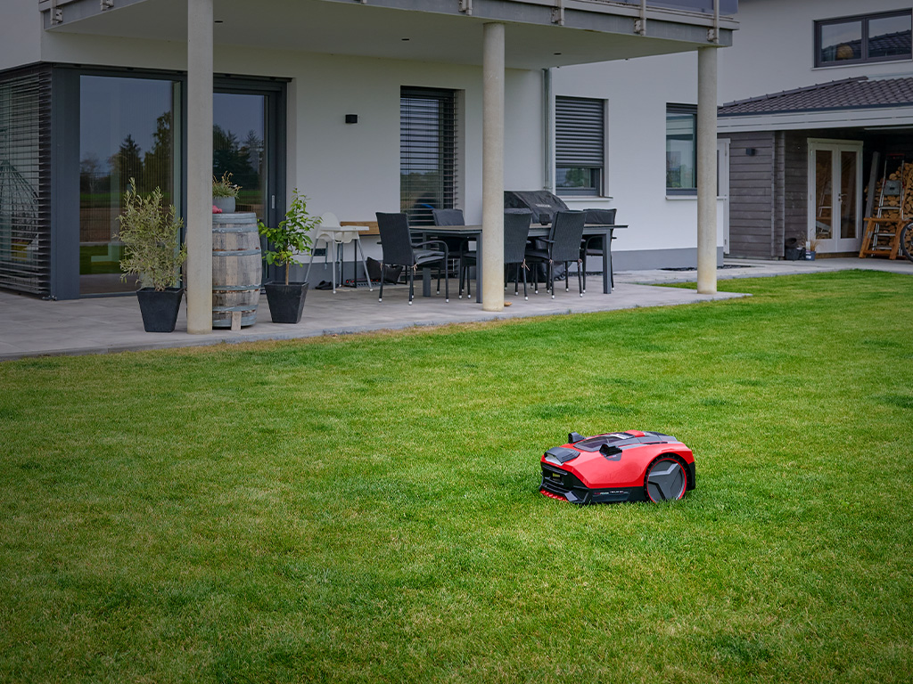 The Einhell robotic lawn mower FREELEXO CAM drives across a well-kept lawn in front of a modern house.