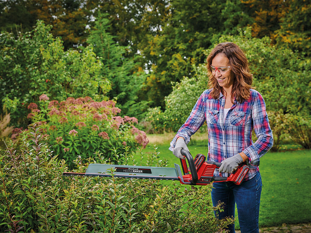 A woman cutting a hedge with an Einhell cordless hedge trimmer.