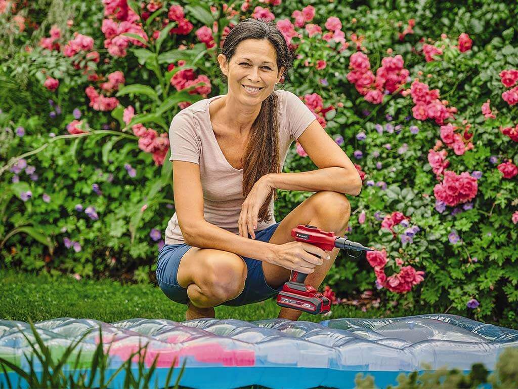 A woman kneels in front of an inflated air mattress, holding the Einhell cordless air pump in one hand.
