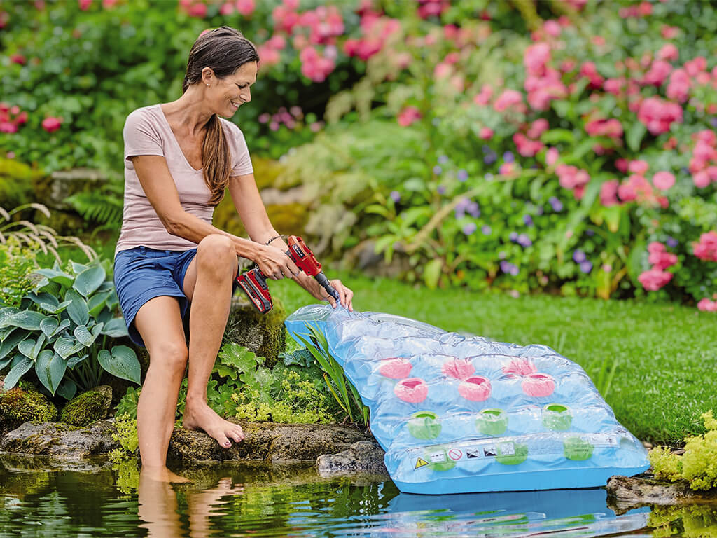 A woman sits next to a pond in her garden and pumps up an air mattress with a cordless Einhell compressor.