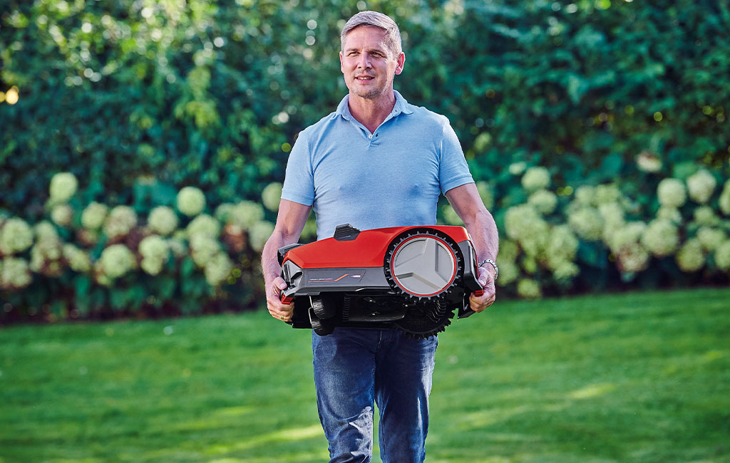 Ein Mann trägt einen roten Mähroboter durch den Garten.