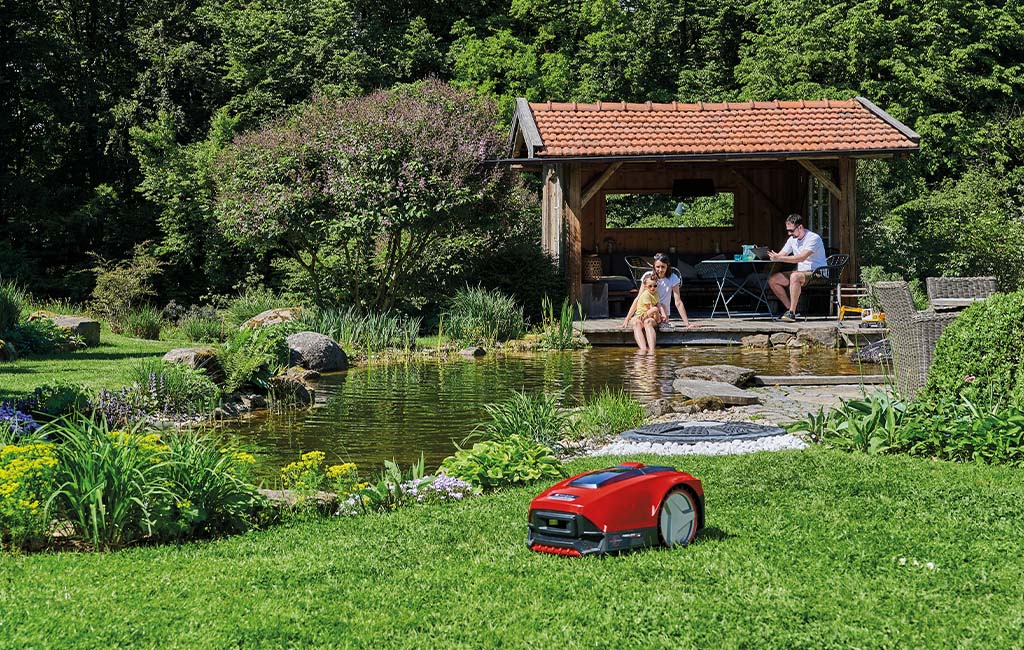 Ein Mähroboter mäht den Rasen in einem Garten, im Hintergrund ein Teich.