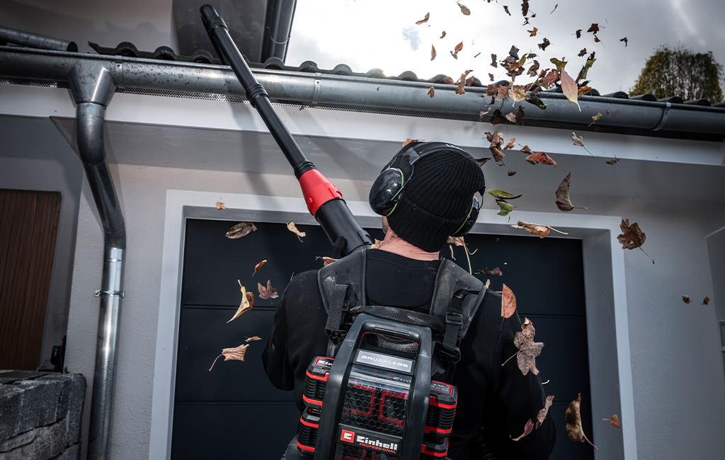 A man uses an Einhell cordless leaf blower with a gutter cleaning attachment to clear a gutter. Leaves fly through the air.